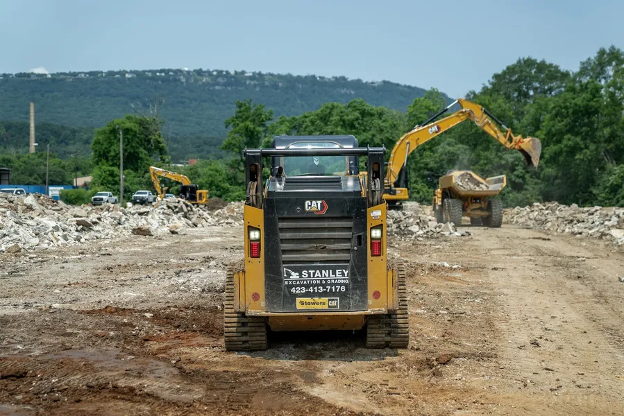 Yellow bulldozer on a brown mound of earth under a blue sky with clouds; another bulldozer in background.